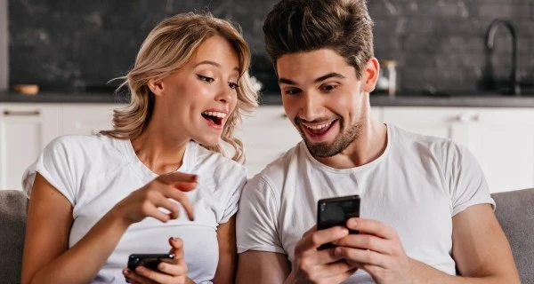 Lächelndes-Paar-sitzt-auf-der-Couch-mit-Handys-–-bezaubernde-junge-Frau-hält-Smartphone-in-der-Hand