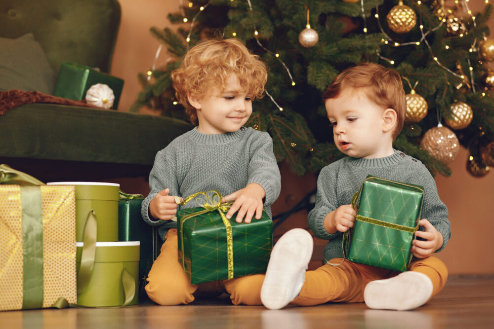 Kleine Kinder in grauen Pullovern neben einem Weihnachtsbaum.